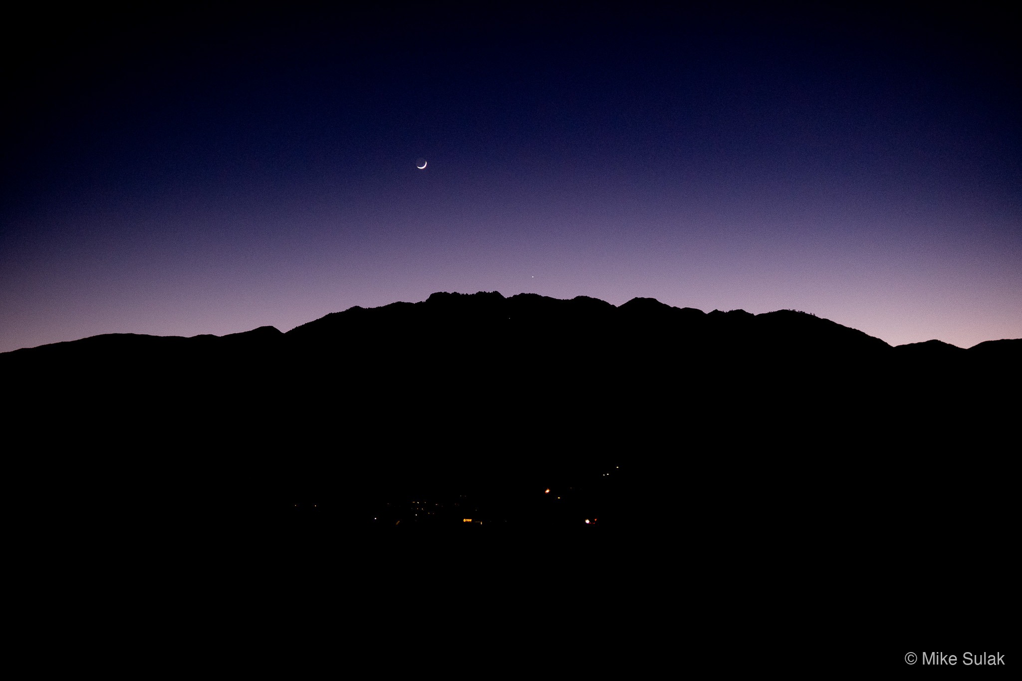 Mountain ridge silhouetted against a deep blue dusk sky with a crescent moon