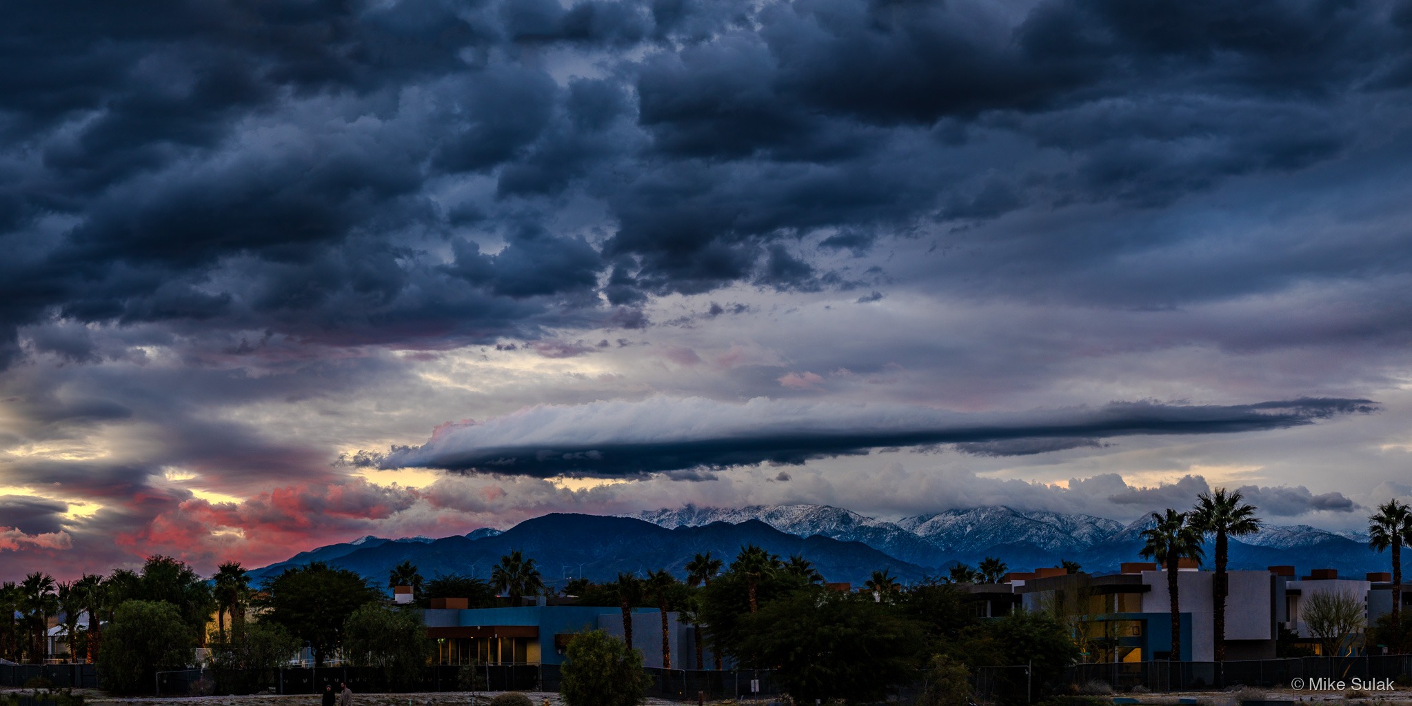 Wide panorama of a desert city at sunset beneath dramatic storm clouds and snow-capped mountains